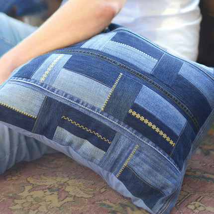 A rectangular throw pillow made from blue denim patches with decorative gold-colored stitching, held by a person sitting on a patterned carpet.