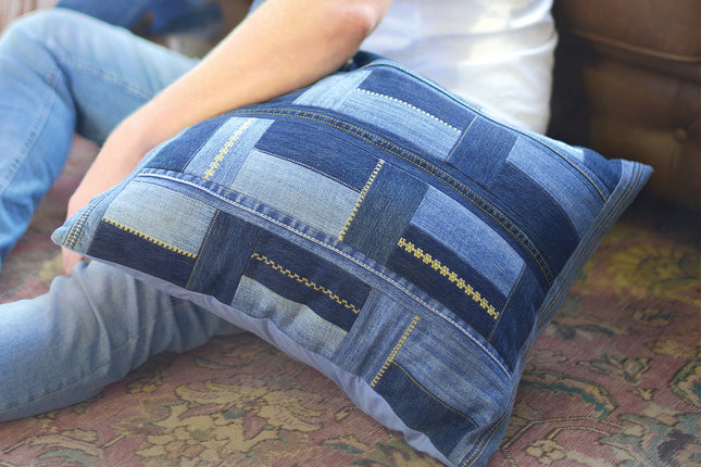 A rectangular throw pillow made from blue denim patches with decorative gold-colored stitching, held by a person sitting on a patterned carpet.