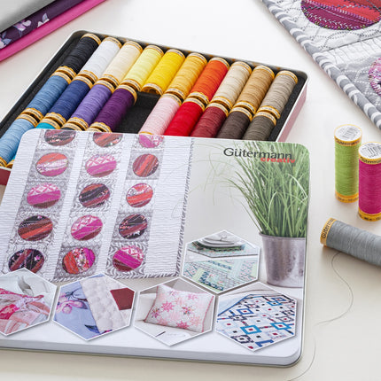 A Gütermann sewing thread tin set displayed on a light surface, featuring a large printed tin cover, an open metal tin filled with colorful thread spools, two loose thread spools, and a quilted fabric sample in the background.
