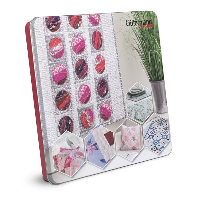 A Gütermann cotton thread tin standing upright at an angle, showing a cover with various textile swatches, circular patterned samples, and small hexagonal inset photos of fabrics and home textiles. The tin has a red spine edge and a white cover with printed textile imagery.