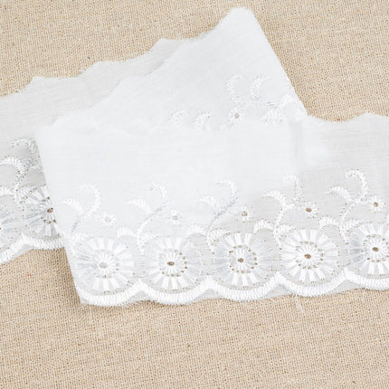 White floral lace trim with scalloped edges and embroidered details, laid flat on a beige textured fabric background.